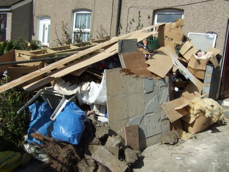 A large pile of mixed household waste and building debris is situated on a residential pavement outside a row of beige-colored houses in Earls Court. The heap includes broken concrete blocks, wooden planks, cardboard boxes, and various discarded materials. A blue plastic bag and a white fabric sack are visible among the rubble, with some cardboard packaging leaning against the concrete debris. The objects are haphazardly stacked, partially covering the pavement and extending towards the front garden of the houses. The background shows a typical London residential setting with windows and curtains visible in the upper floors of the buildings. The scene is illuminated by daylight, casting soft shadows on the rubbish. This image illustrates rubbish accumulation suitable for removal services provided by Office Clearance Earls Court, highlighting the type of waste commonly managed in the area.