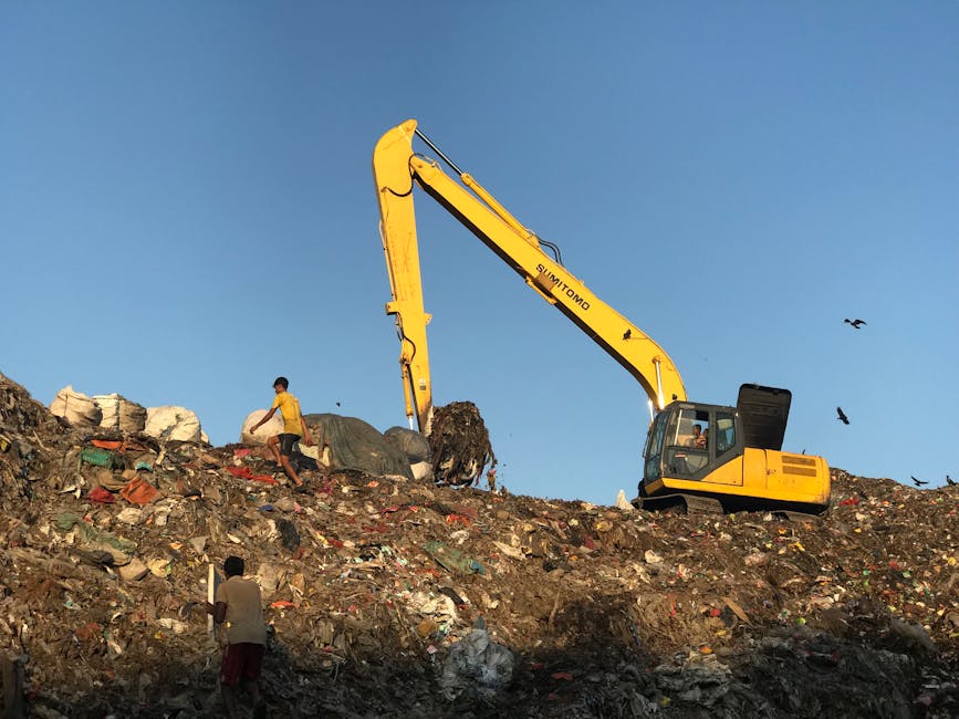 A large pile of mixed waste materials is situated on a flat asphalt surface, with a blue-painted metal structure on the left side of the image. The waste consists primarily of crushed or broken plasti