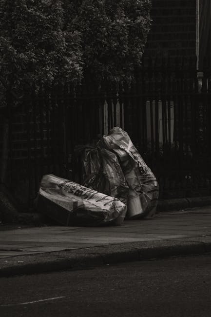 Two large black plastic rubbish bags filled with waste are placed on the edge of a sidewalk near a black metal fence. The bags appear to contain household or construction debris, with some partially t