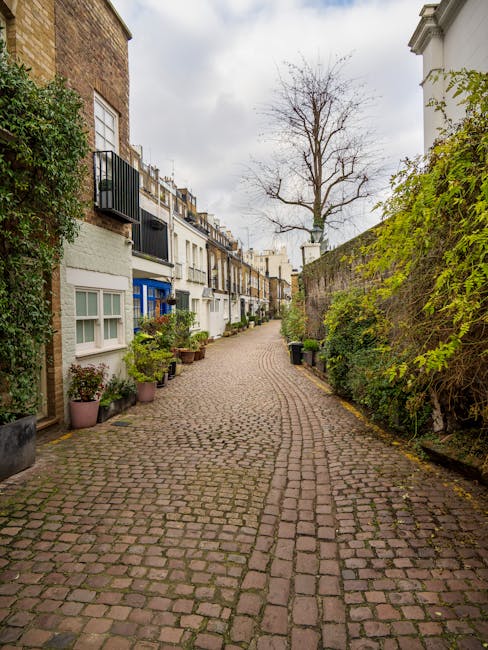 A narrow cobblestone alleyway in a residential area, lined with terraced houses on the left and a tall, leafy brick wall on the right, with various potted plants and small shrubs along the base of the